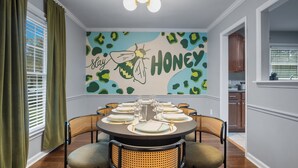 A dining area with a round table set for six, surrounded by wicker chairs. A wall mural with a bee and the words "Stay Honey" is visible. - 8 Beds, Large Groups, Karaoke, Firepit  (Charlotte)