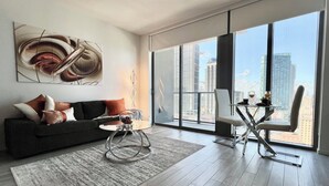 A modern living room with a dark sofa, a glass coffee table, and a view of tall buildings through large windows. - Lux apartment in Miami downtown (Miami)