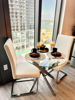 A modern dining area with a glass table, two white cushioned chairs, and a view of a beach and high-rise buildings. - Lux apartment in Miami downtown (Miami)