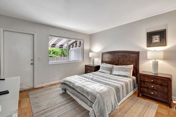 Une chambre avec un grand lit, deux tables de chevet avec des lampes, une fenêtre donnant sur un paysage verdoyant et une porte.
