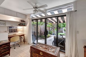 Une pièce comprenant un bureau, une chaise et un coffre en bois décoratif. On y trouve également un ventilateur de plafond, une fenêtre avec des rideaux et une vue sur un vélo à l’extérieur.