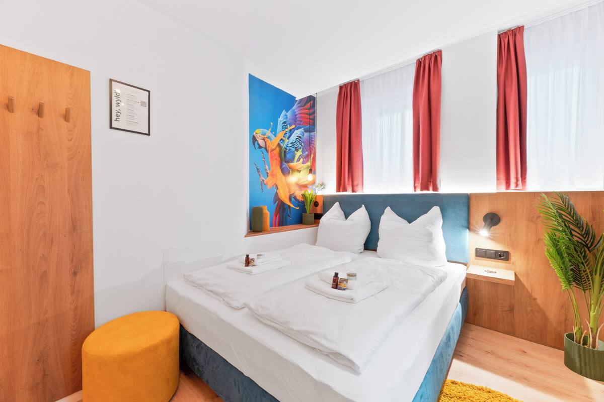 Une chambre d’hôtel moderne avec un grand lit, deux tables de chevet, une tête de lit en bois, une œuvre d’art accrochée au mur et une fenêtre avec des rideaux rouges.