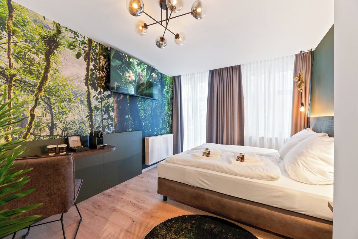 Une chambre d’hôtel moderne dotée d’un grand lit, d’un bureau avec un ordinateur, d’un téléviseur fixé au mur et d’une fresque murale sur le thème de la forêt.