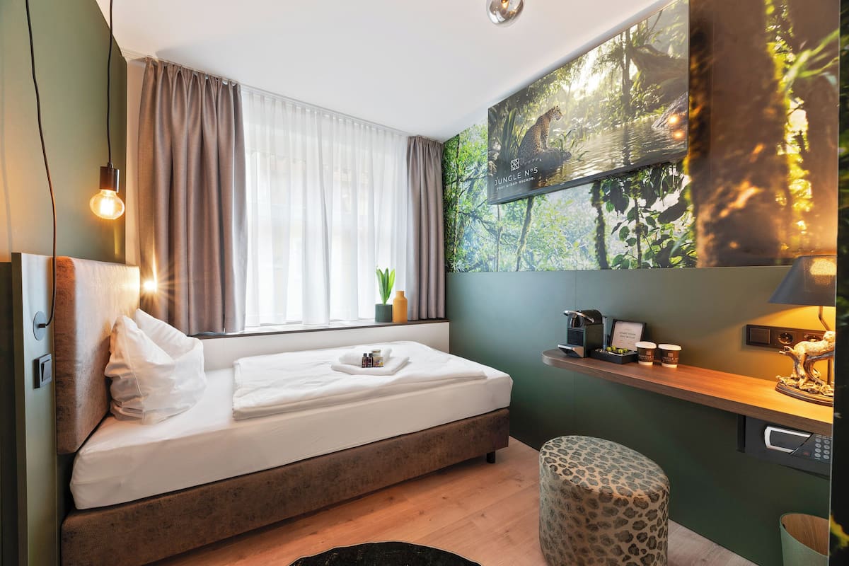 Une chambre moderne avec un lit, une fenêtre avec des rideaux, un tableau représentant une scène de jungle accroché au mur, une petite table avec une lampe et un banc.
