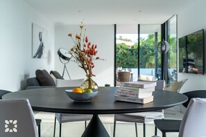 A modern dining area with a round table, a vase with dried flowers, and a bowl of fruit.