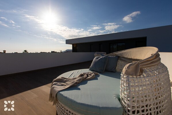 Terraza en la azotea con zona de estar que incluye un sofá con cojines azul claro, una manta beige y una silla blanca trenzada.