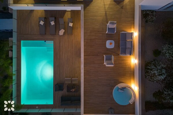 Vista aérea de un moderno espacio exterior con piscina, terraza de madera, sillas de descanso y una zona de estar circular.