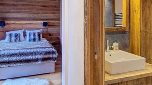 Een houten slaapkamer met een bed, kussens en een zacht, wit vloerkleed. Er staat een houten kast met een wastafel en een spiegel erboven.