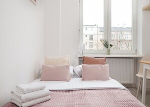 Un dormitorio con cama, almohadas, una mesita de noche, una silla y una ventana con vista a edificios.