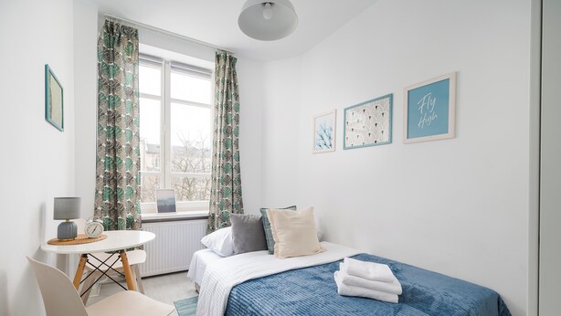 Un dormitorio ordenado con cama, una mesita de noche con lámpara, una silla y una ventana con cortinas.