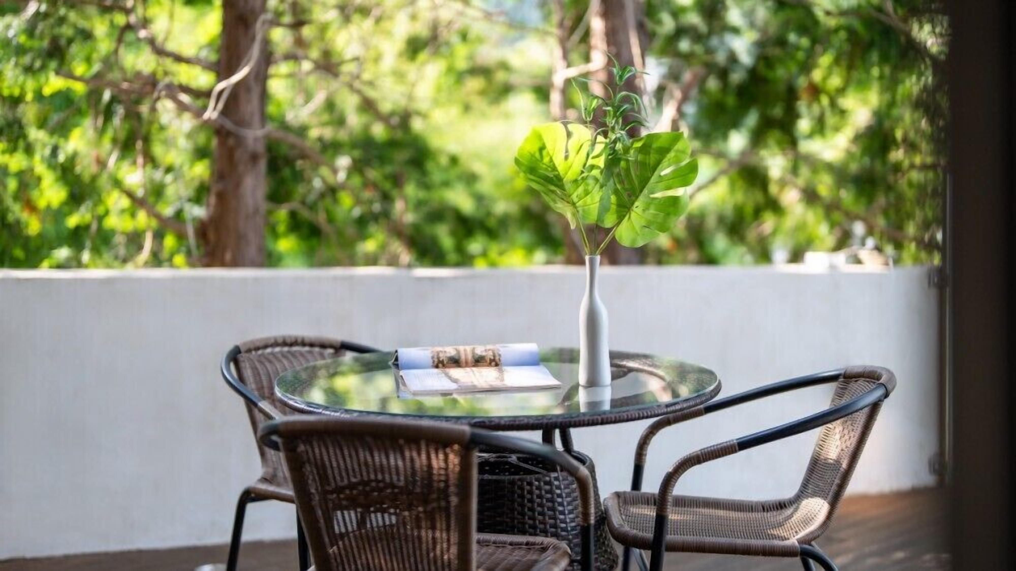 A round glass table with a white vase and a rolled-up menu, surrounded by wicker chairs on a wooden deck with a white wall and greenery in the background.