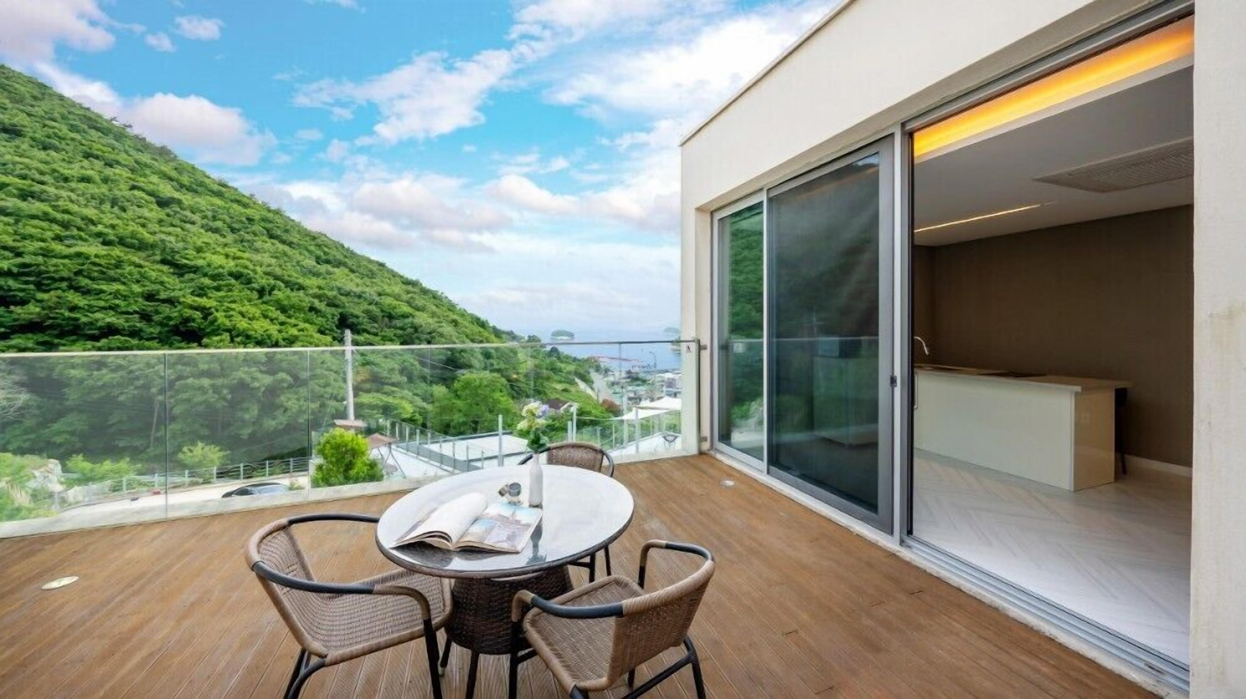 Un balcon avec une table ronde et des chaises, une porte vitrée donnant sur un espace intérieur, et une vue sur une colline verdoyante et une étendue d’eau.