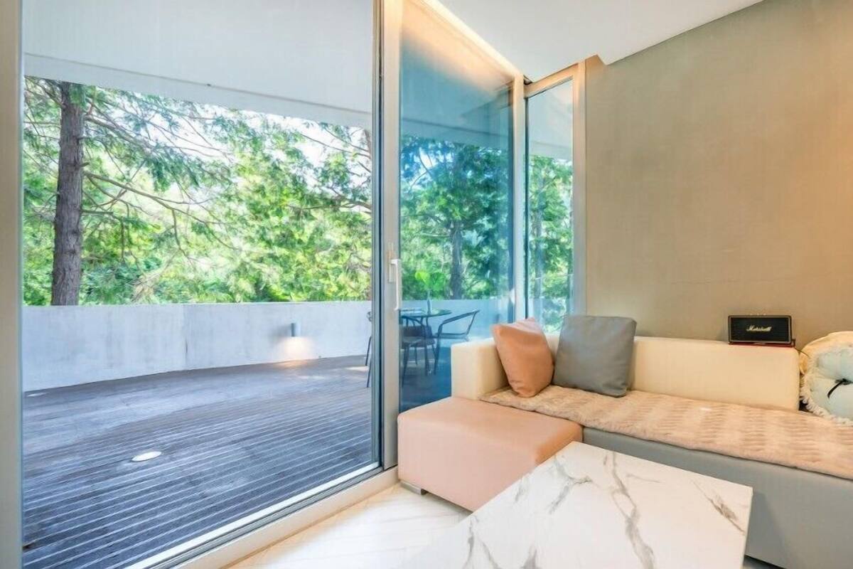 A modern living room with a glass wall, a sofa with cushions, a marble coffee table, and a small speaker.