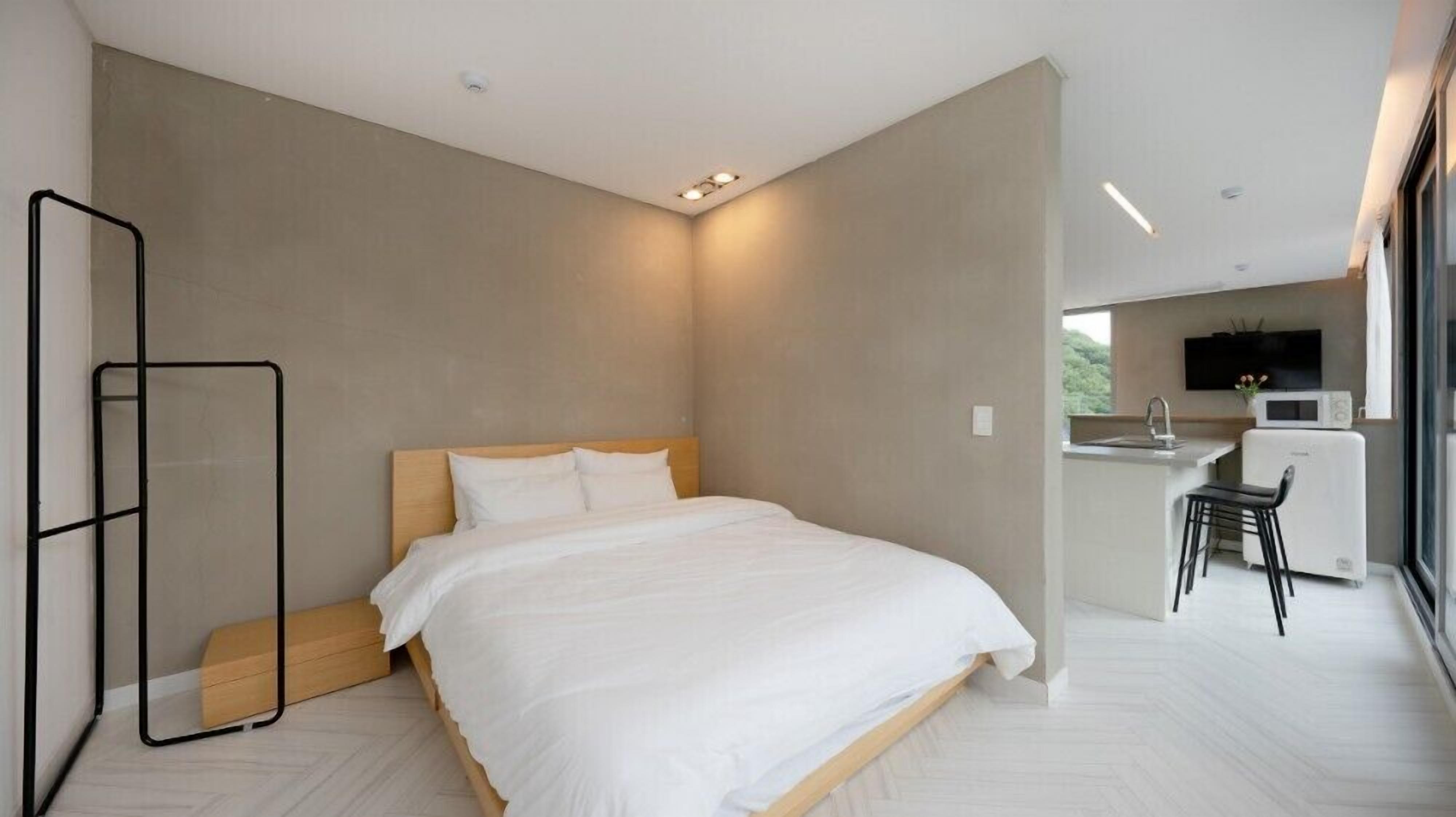 A modern bedroom with a large bed, a small kitchenette, and a view of greenery outside.