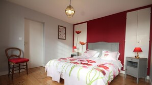 Un dormitorio con suelo de madera, una cama con ropa de cama floral, una silla con un cojín rojo, una mesita de noche con una lámpara y una pared con un cuadro enmarcado.
