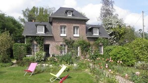 Una casa de ladrillo de dos pisos con jardín, tres sillas de jardín y flores floreciendo.
