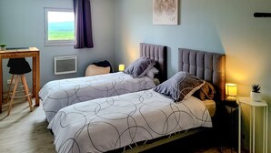 A bedroom with two beds, a desk, a chair, a radiator, and a window with curtains.
