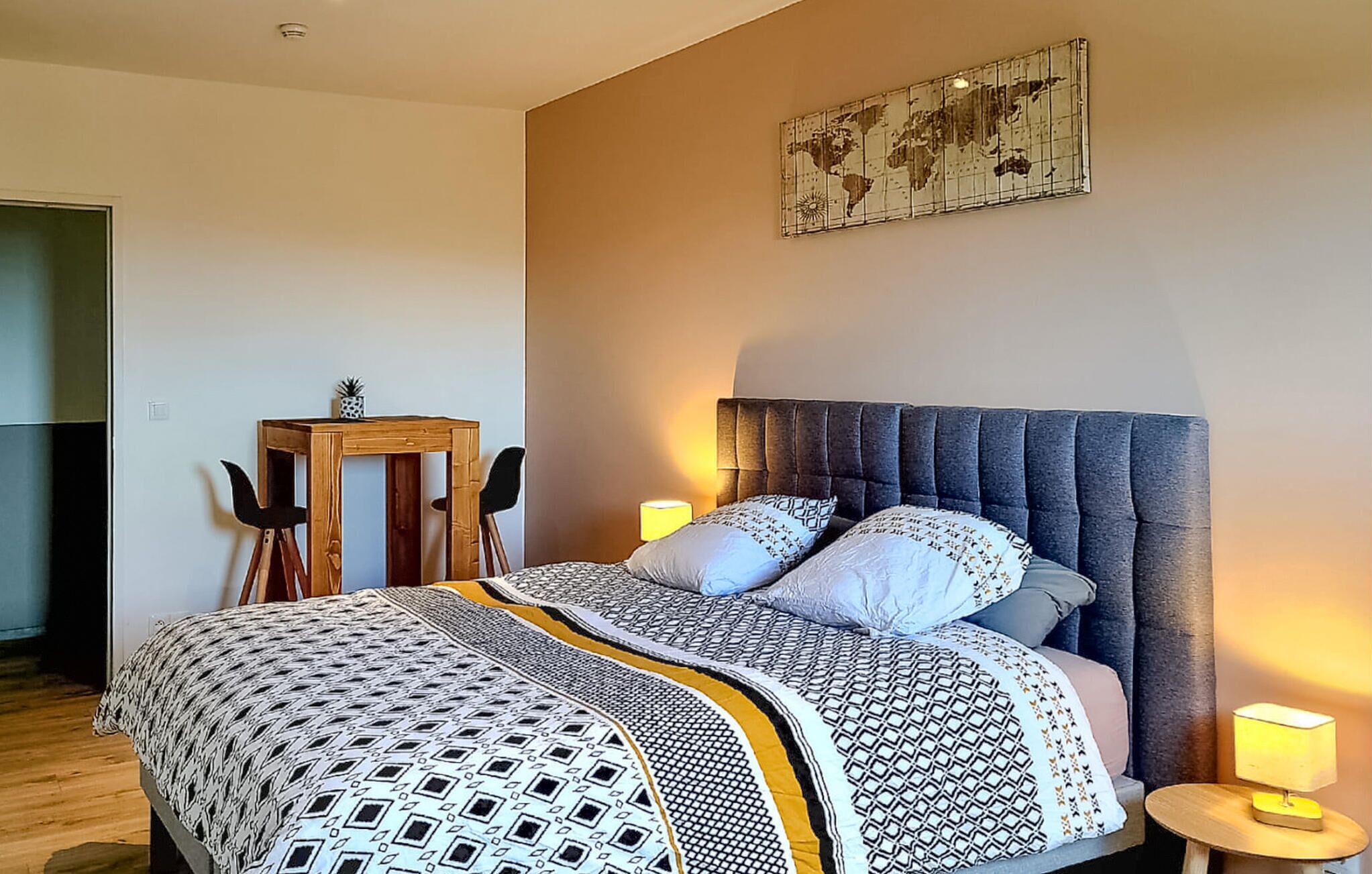 Una camera da letto con un letto, due lampade da comodino, un piccolo tavolo di legno con una sedia e un'opera d'arte raffigurante una mappa del mondo sulla parete.