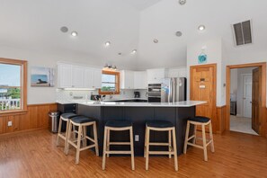 Una cocina moderna con armarios blancos, una isla central con taburetes, electrodomésticos de acero inoxidable y vistas al exterior.