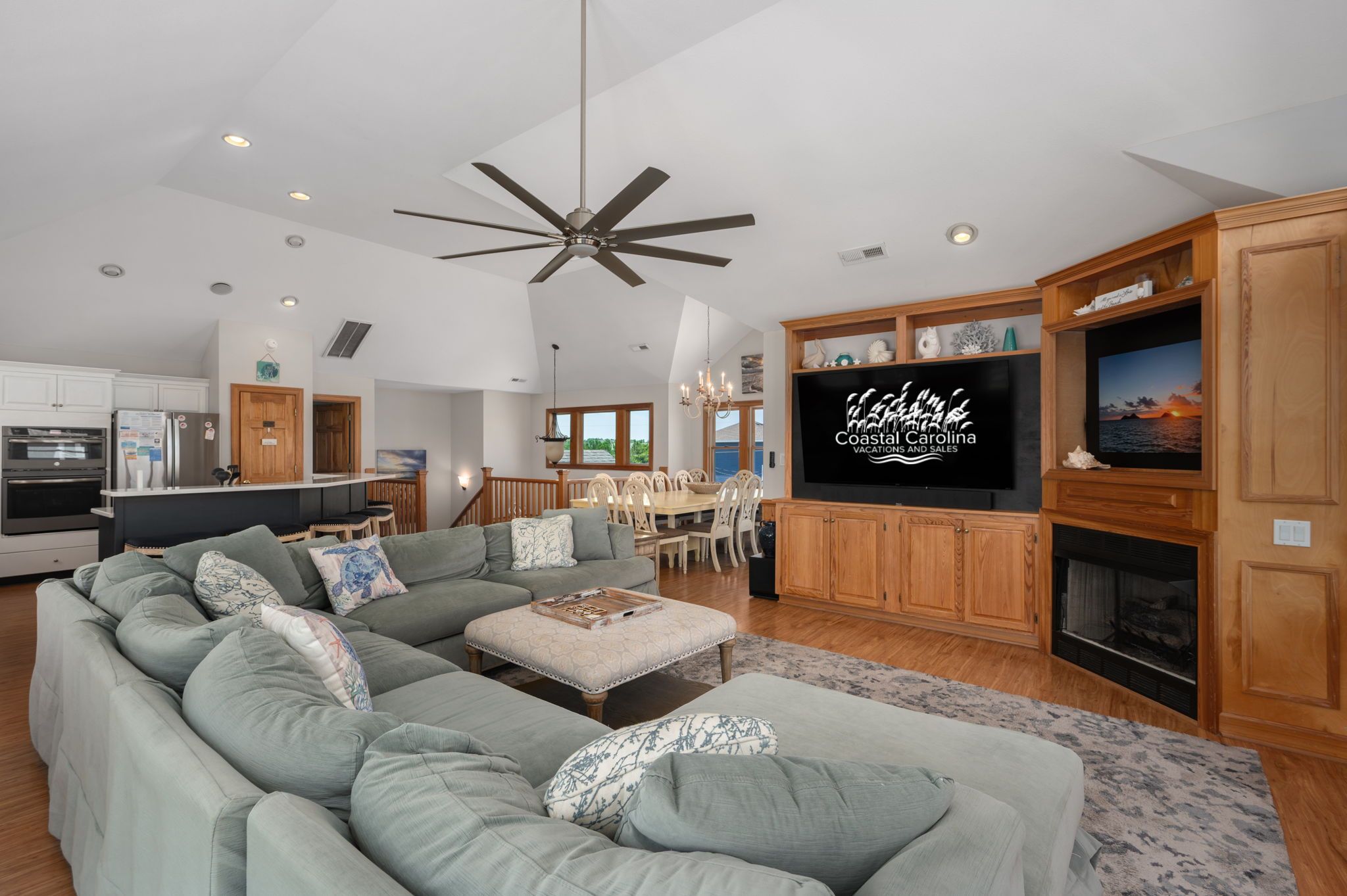 Ein großzügiges Wohnzimmer mit einer großen Eckcouch, einem Essbereich, einem Kamin und einem Fernseher, der 'Coastal Carolina Vacations and Sales' anzeigt.