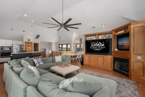 Amplia sala de estar con un sofá modular grande, comedor, chimenea y un televisor mostrando 'Coastal Carolina Vacations and Sales'.