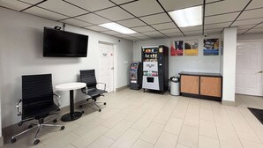 A waiting area with two black office chairs, a small round table, a vending machine, a trash can, and a reception desk. - Studio 6 Sulphur, LA (Sulphur)