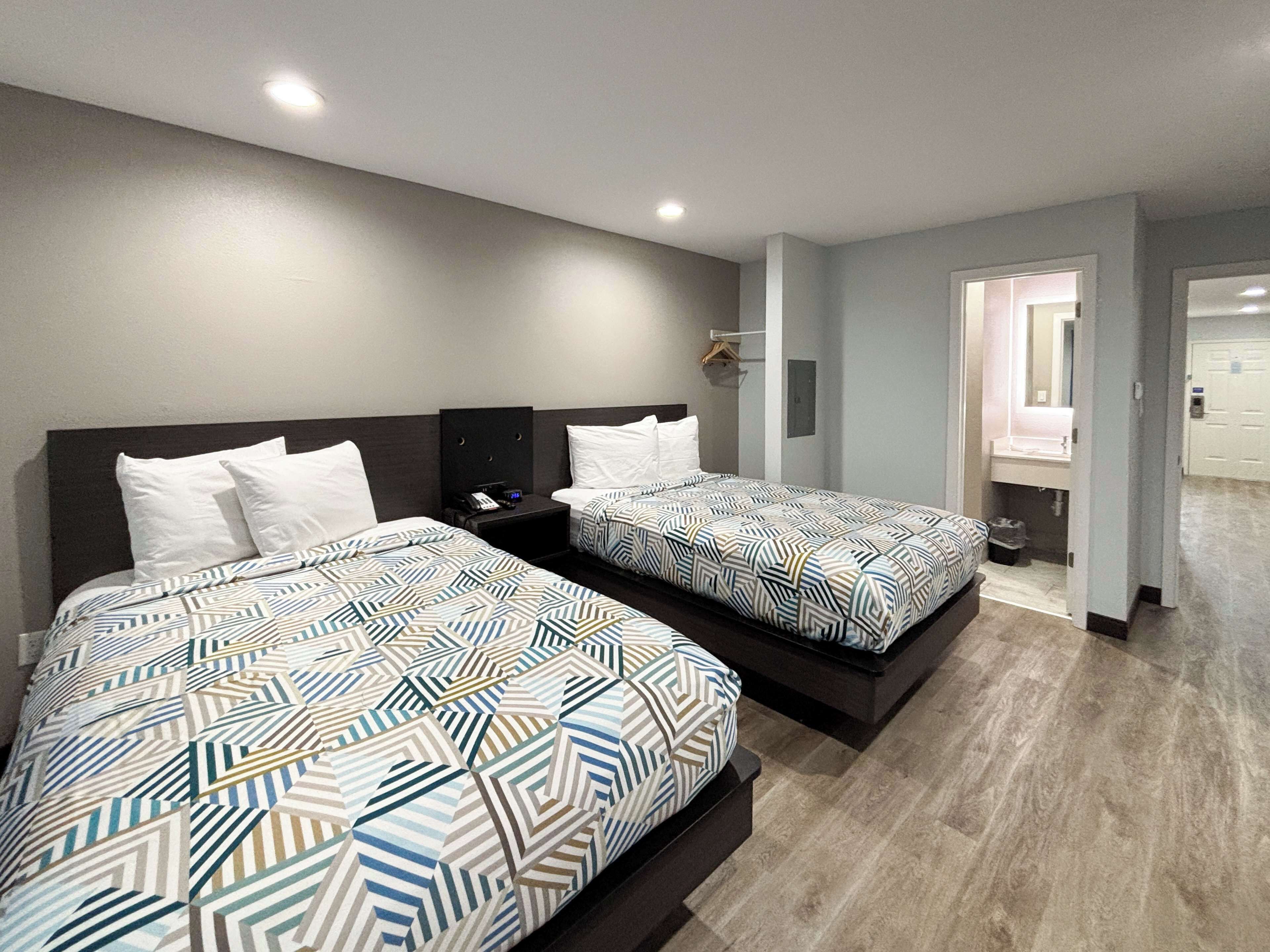 A hotel room with two beds, each with a patterned bedspread, a bedside table with a phone, and a wall-mounted light.