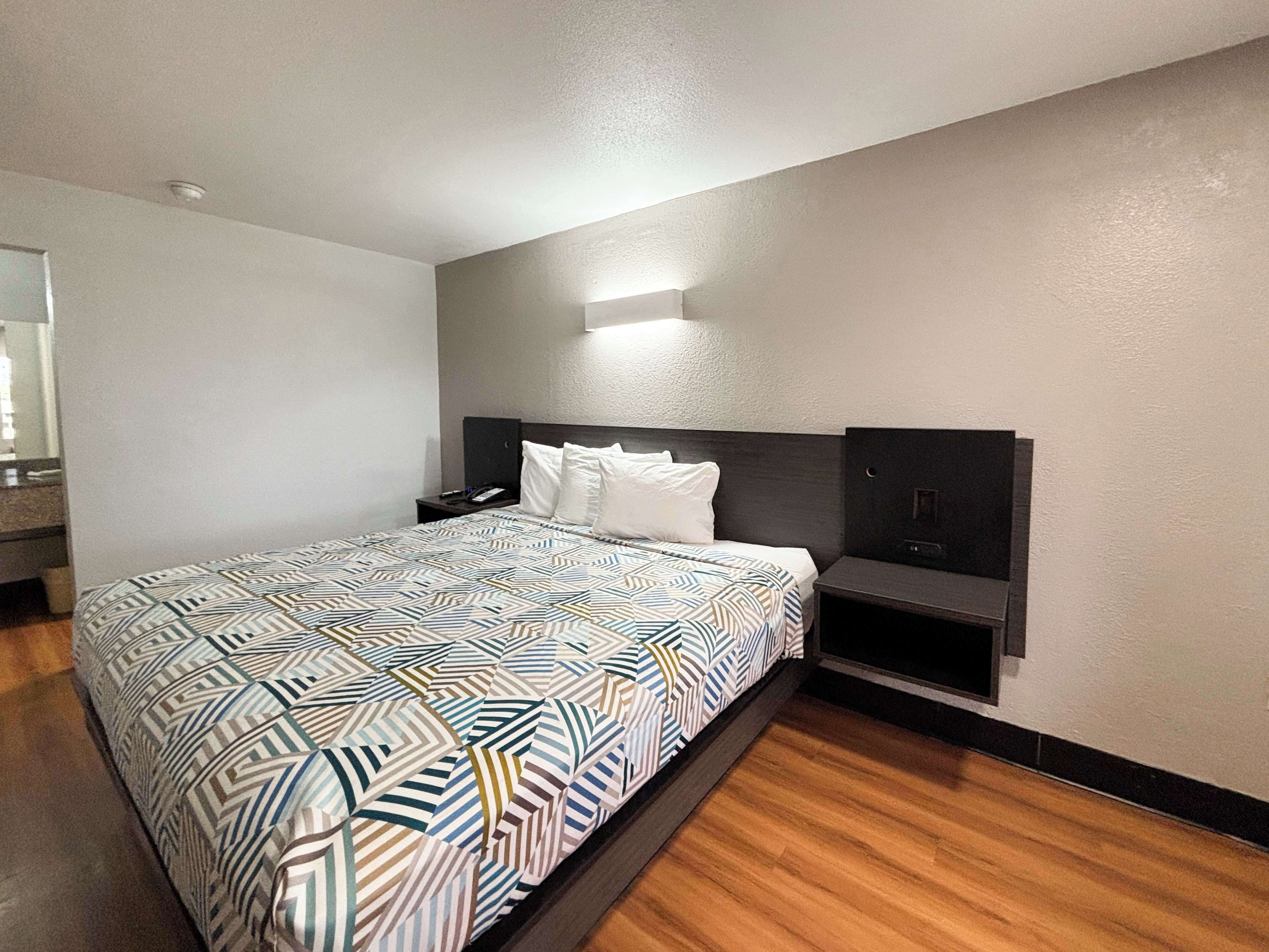 Habitación de hotel con una cama grande, una mesita de noche, una lámpara y un televisor montado en la pared, además de un espejo.