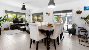A modern dining area with a wooden table, white upholstered chairs, and a television on a dark wooden cabinet. - The Beach Barn - Balnarring Beach (Balnarring Beach)