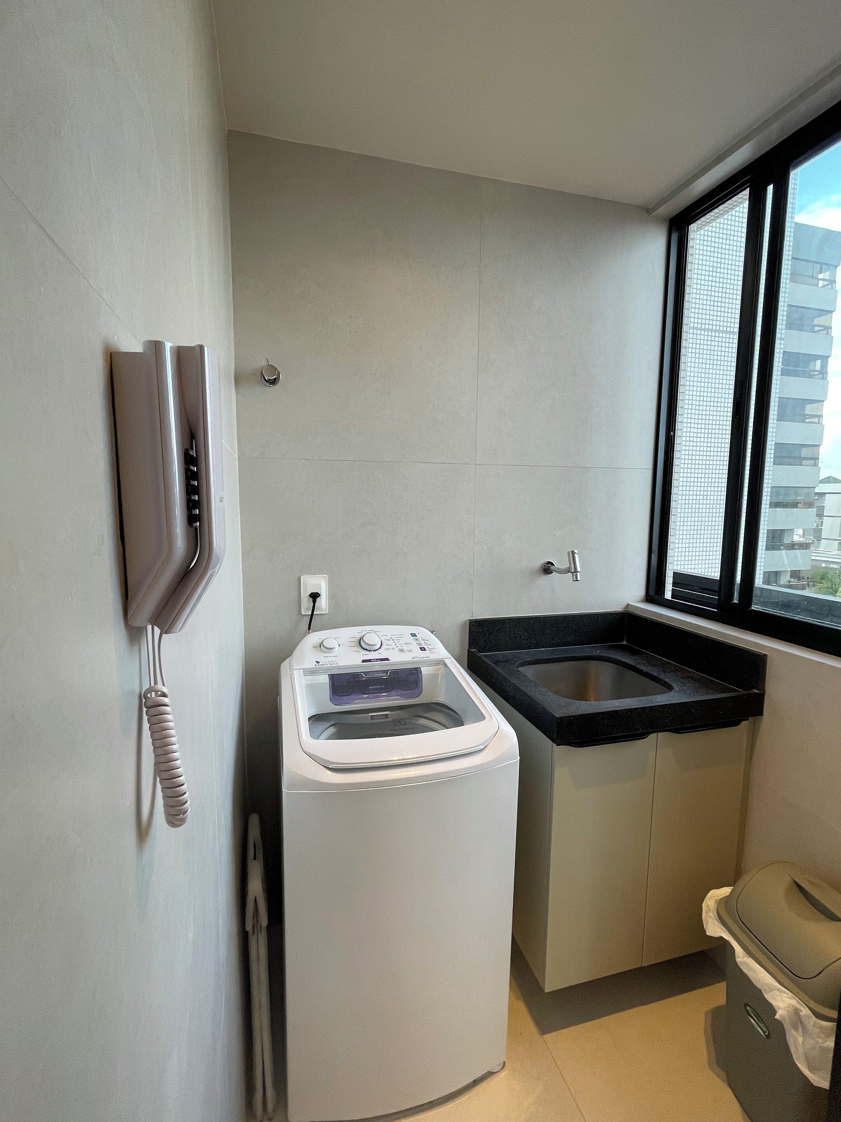 A compact laundry room with a front-loading washing machine, a sink, and a trash bin.