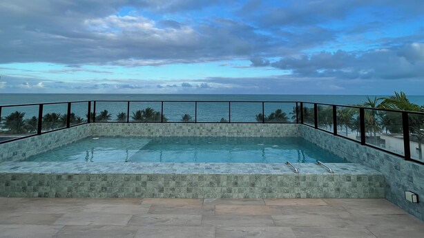 A rectangular pool with a stone border, surrounded by a black railing, overlooking a beach with palm trees and the ocean under a partly cloudy sky.