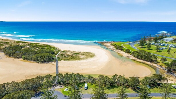A coastal area with a sandy beach, a clear blue ocean, and a parking lot with vehicles.