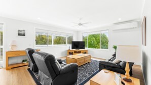 A modern living room with a flat-screen TV, leather sofas, a wooden coffee table, and large windows offering a view of trees.