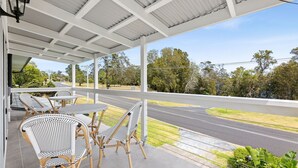 A covered outdoor seating area with wicker chairs and tables, overlooking a road and greenery.