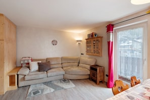 A living room with a curved sofa, a small wooden cabinet, a table with chairs, and a window with a curtain. - Apartment 'Giulia' with Mountain View, Shared Garden and Balcony (Canazei)
