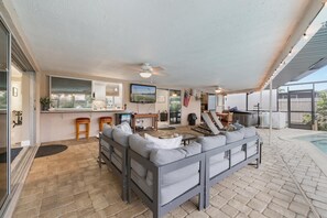 A covered patio area with a seating arrangement, a television, and a pool in the background.
