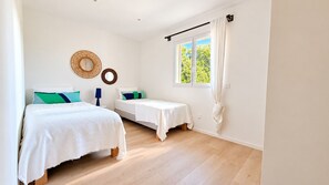 A bedroom with two beds, a window with curtains, a wall-mounted mirror, and a wooden floor.