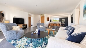 A modern living room with a sofa, armchair, and coffee table. There is a television, a dining area, and a kitchen in the background.