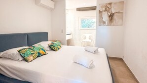 Un lit bien fait, avec du linge de lit blanc et des coussins aux motifs tropicaux, une tête de lit bleue et un tableau accroché au mur.