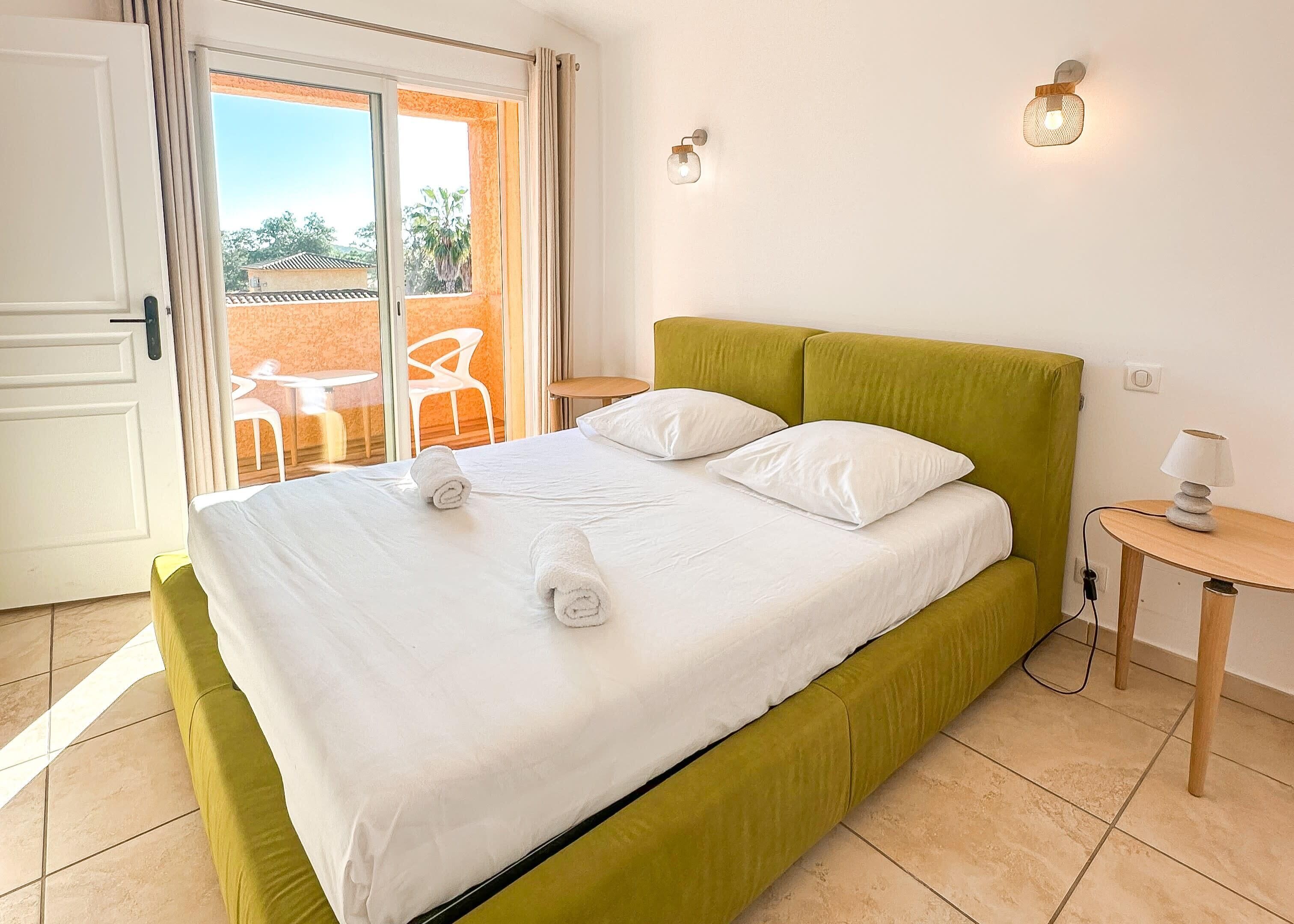A neatly made bed with white linens and two rolled towels, a green upholstered headboard, a small bedside table with a lamp, and a view of an outdoor patio with chairs and a table through a glass door.