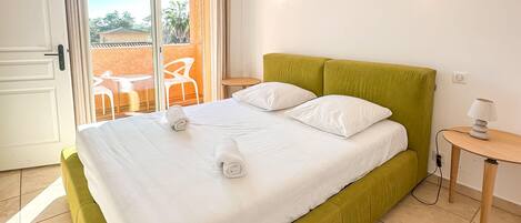 A neatly made bed with white linens and two rolled towels, a green upholstered headboard, a small bedside table with a lamp, and a view of an outdoor patio with chairs and a table through a glass door.