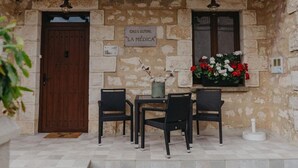 A rustic stone building with a wooden door, a sign reading 'CASA RURAL LA MÉDICA,' outdoor seating with chairs and a table, and a flower box with red and white flowers.