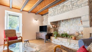 A living room with a stone fireplace, wooden beams, a glass coffee table, and a TV on a wooden cabinet. - Holiday Home 'Les Hirondelles' with Private Terrace, Private Garden and Wi-Fi (Mellac)