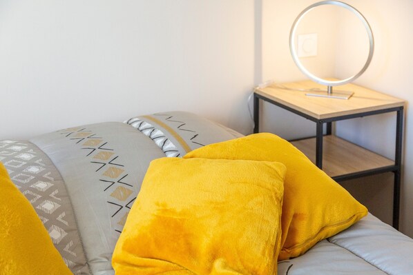 A bed with a patterned bedspread, two pillows, a bedside table with a mirror, and a wall socket.