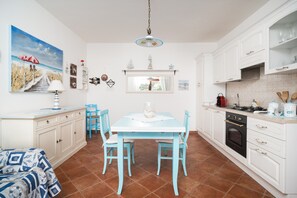 A kitchen with white cabinets, a blue table, and chairs, a dining area with a window, and a painting of a beach scene. - S'ercole Deluxe Apartment with Covered Veranda and Barbecue, Perfect for Couples (Posada)
