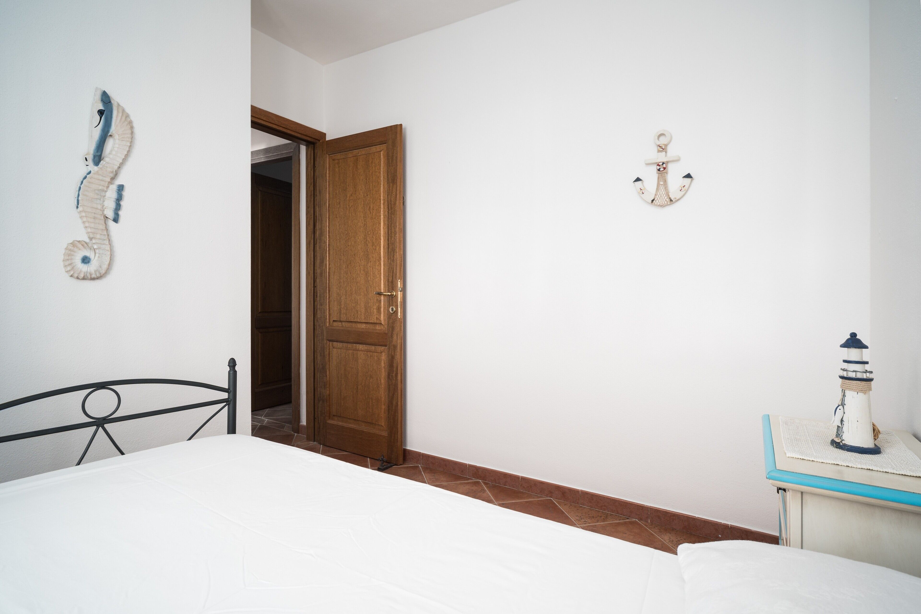 Quarto com cama, porta de madeira, decoração de âncora na parede e uma pequena mesa com modelo de farol.