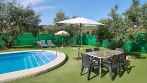 Una piscina con un patio circundante, que incluye una mesa y sillas, y una valla verde al fondo.