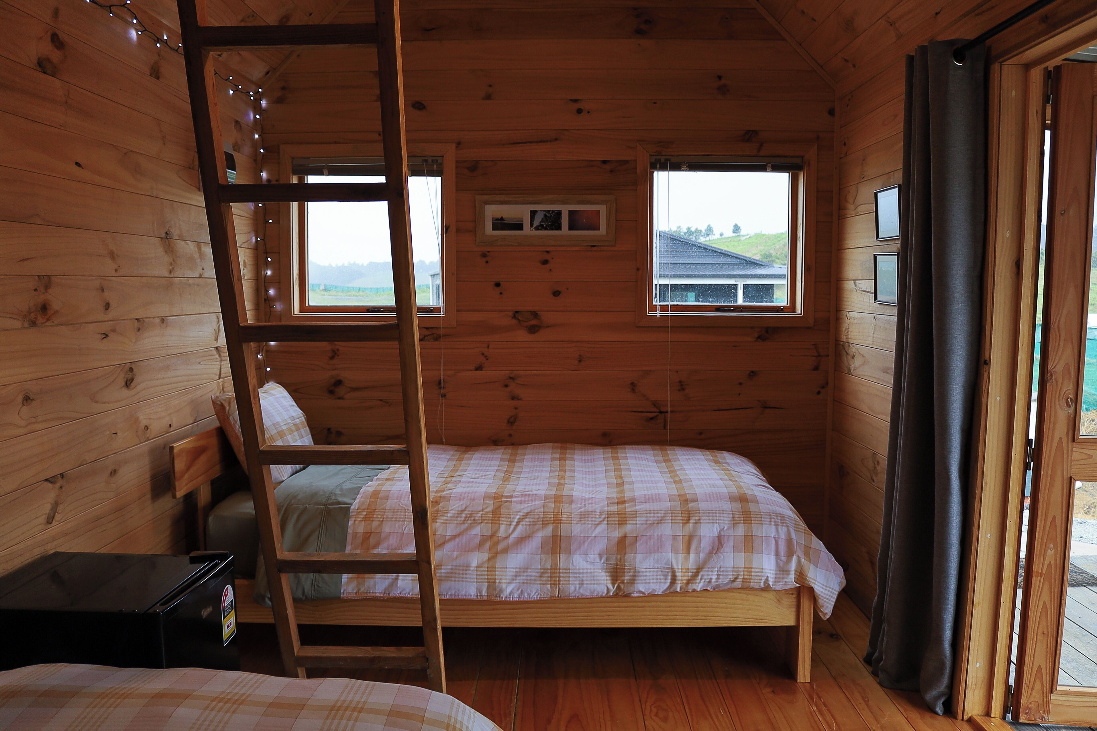 A wooden cabin room with a bunk bed, a ladder, two windows, and a small table.