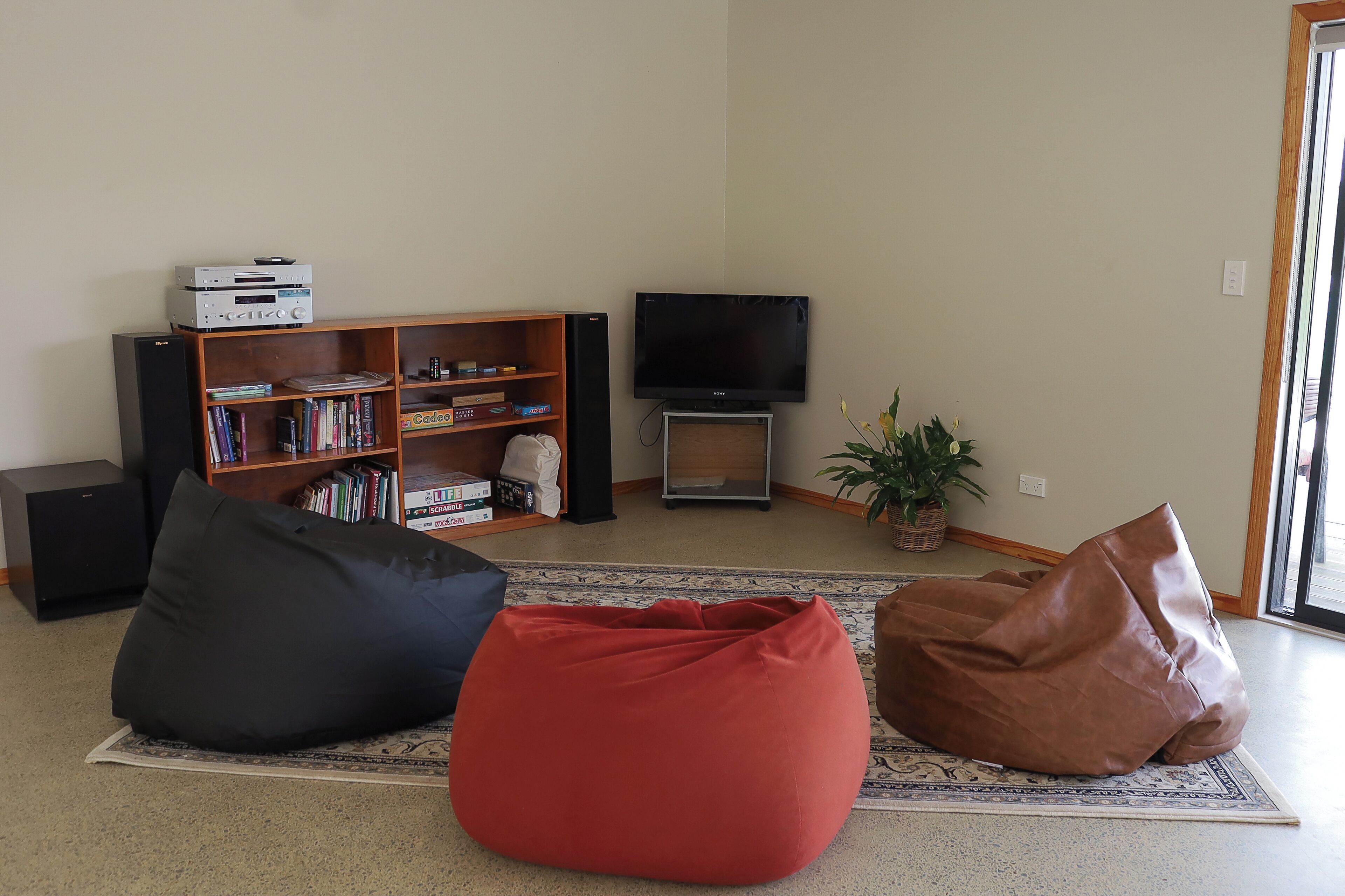 Uma sala de estar com televisão de tela plana, uma estante de livros e dois puffs sobre um tapete estampado.