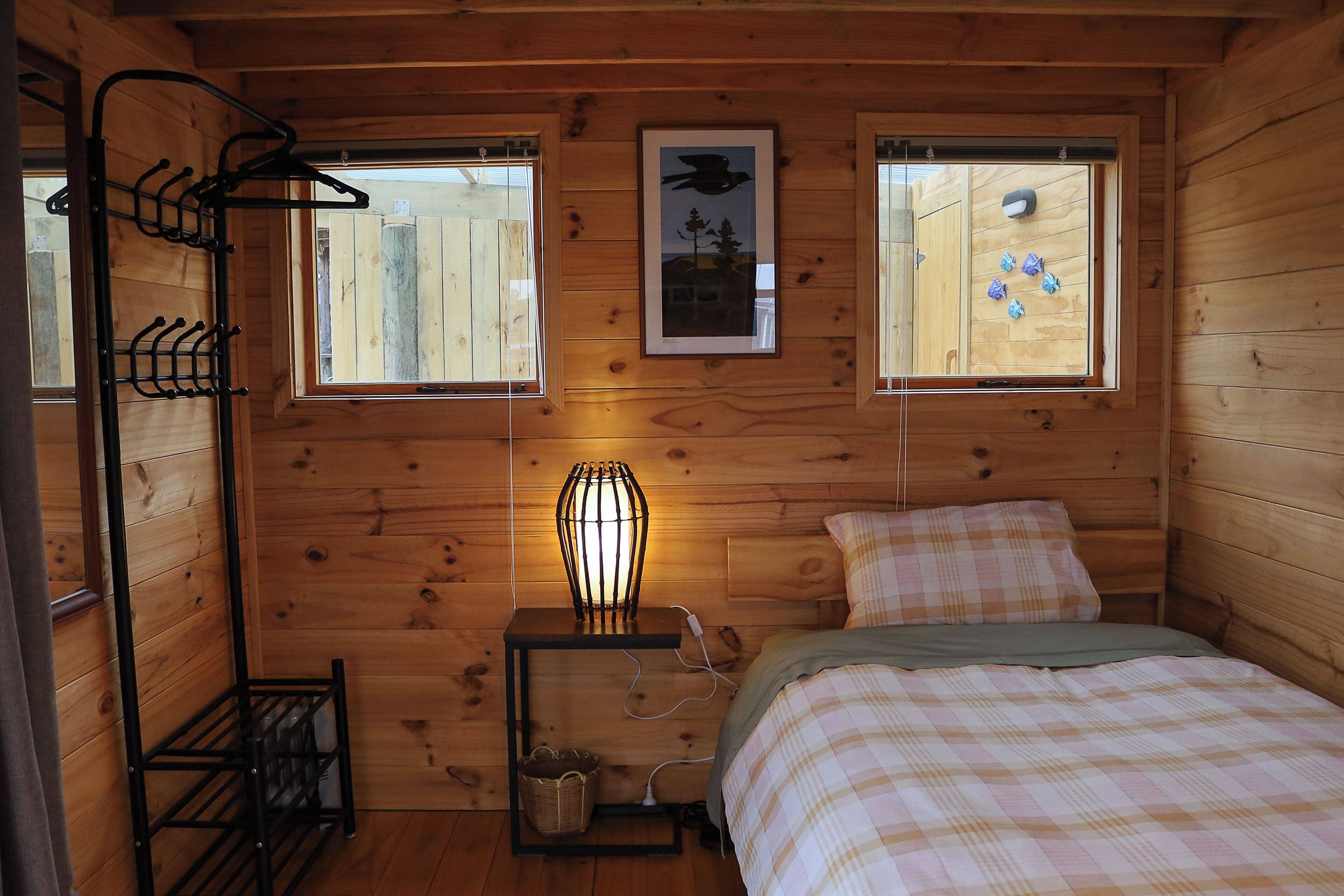 A wooden cabin bedroom with a bed, a hanging clothes rack, a nightstand with a lamp, and two windows.